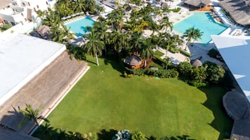 An aerial view of a resort with two swimming pools, palm trees, and thatched-roof structures.