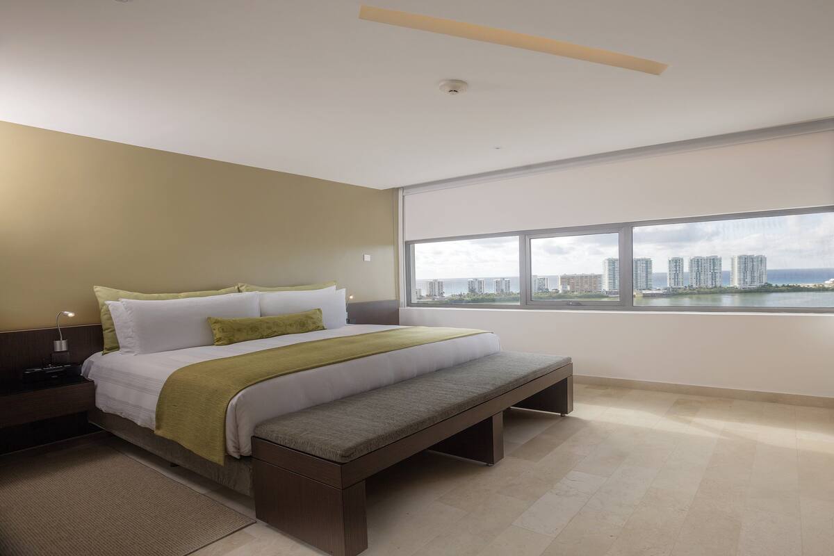 A modern hotel room with a large bed, a bench, a nightstand, and a view of a cityscape through the window.