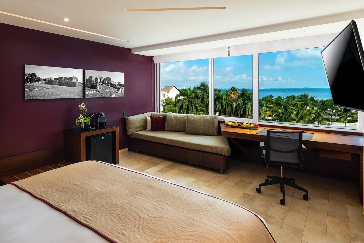 A modern hotel room with a large bed, a sofa, a desk with a computer, and a view of the ocean.