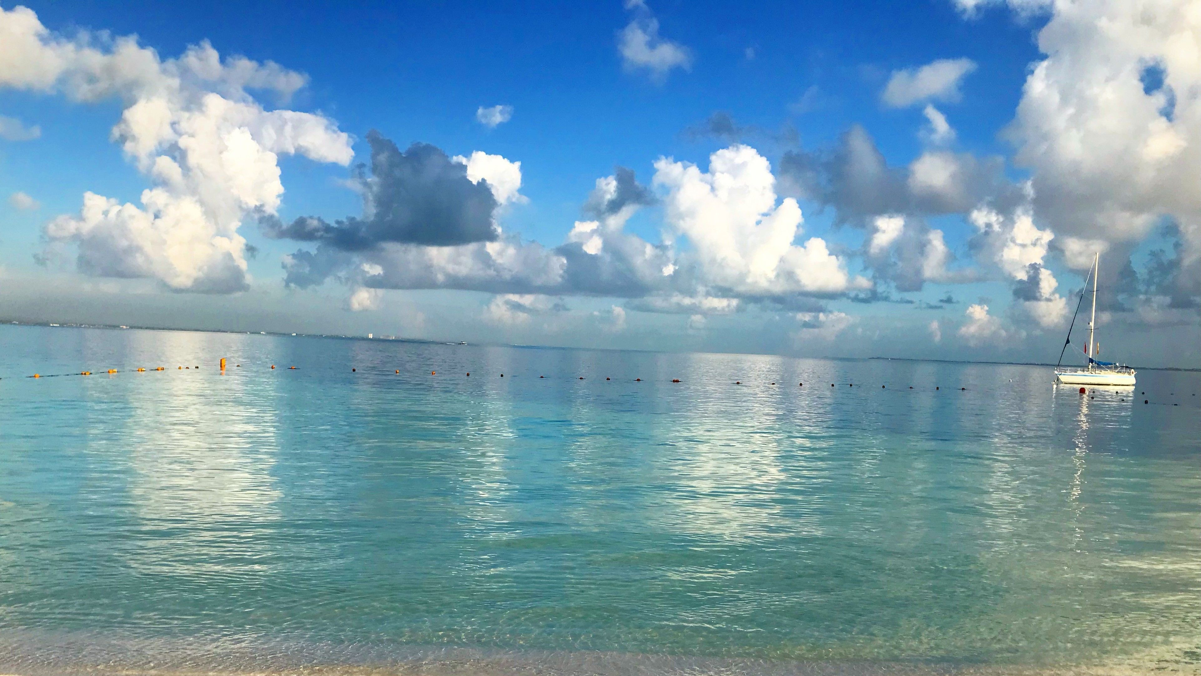 Un mar tranquilo con un velero anclado cerca de la costa, cielos despejados y algunas nubes dispersas.