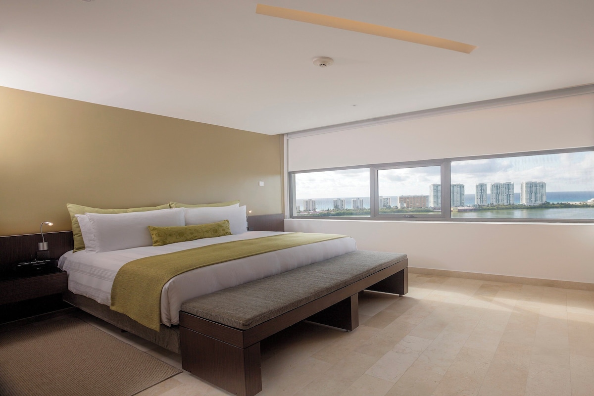 A modern hotel room with a large bed, a bench, a nightstand, and a view of a cityscape through the window.