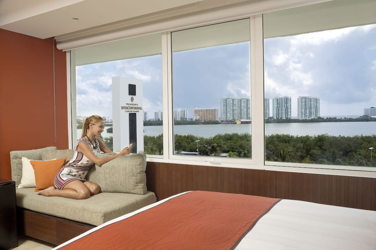 A hotel room with a large window, a bed, a sofa, and a view of a cityscape and water.