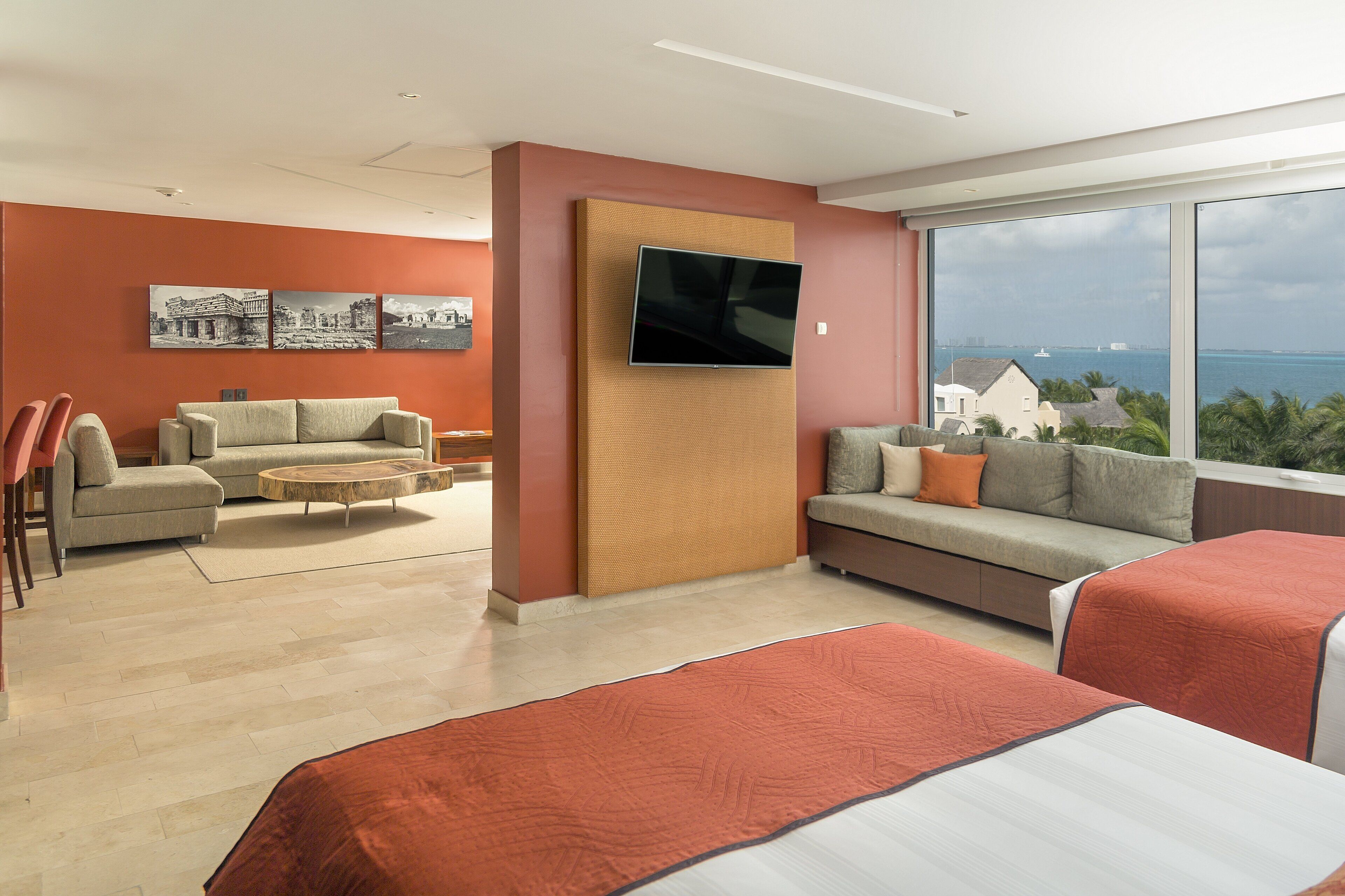 A modern hotel room with a large window offering a view of the sea and buildings, a flat-screen TV mounted on a textured panel, a sofa, a small round coffee table, and two beds with red bedding.