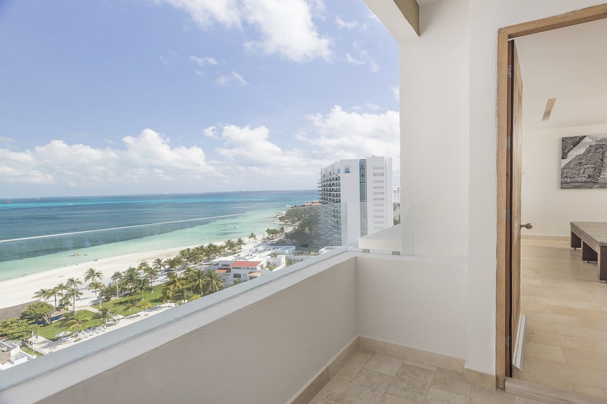 A balcony with a view of a beach and ocean, a bed with a bench, and a painting on the wall.