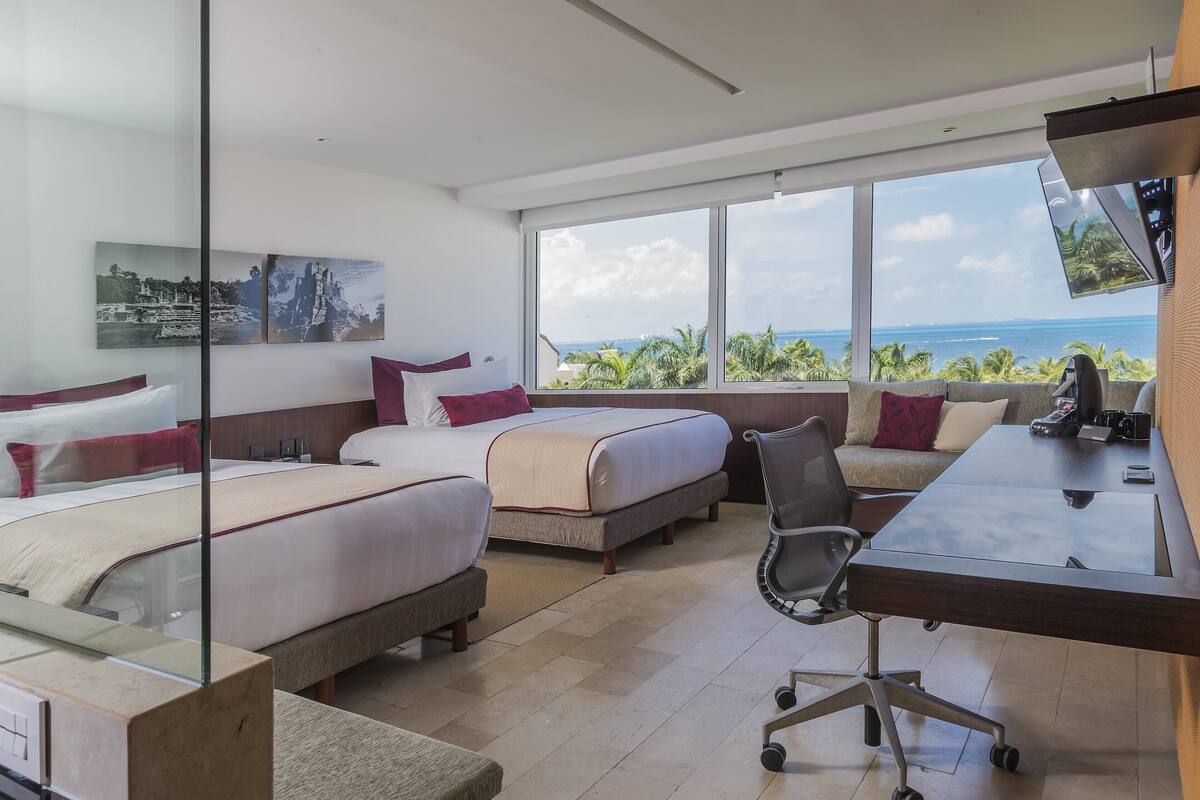 A hotel room with two beds, a desk, and a large window offering a view of the ocean and palm trees.