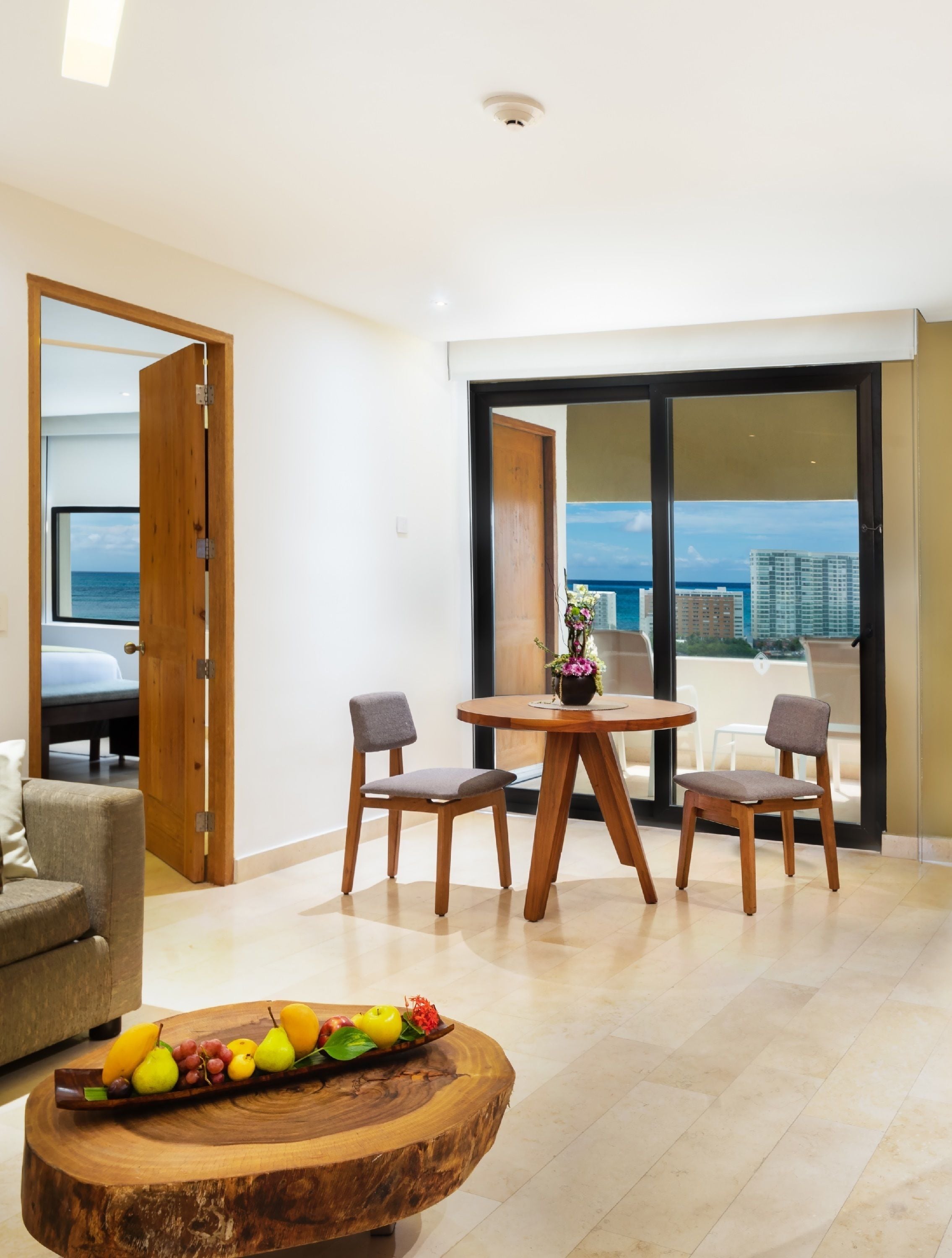 A modern living room with a wooden dining table, two chairs, a sofa, and a coffee table with fruit. A large sliding glass door leads to a balcony with a city view.