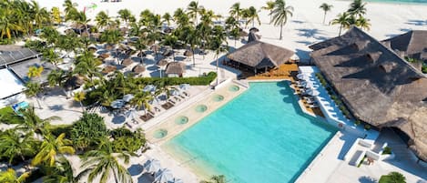 An aerial view of a resort with a large pool, beach, and thatched-roof structures.