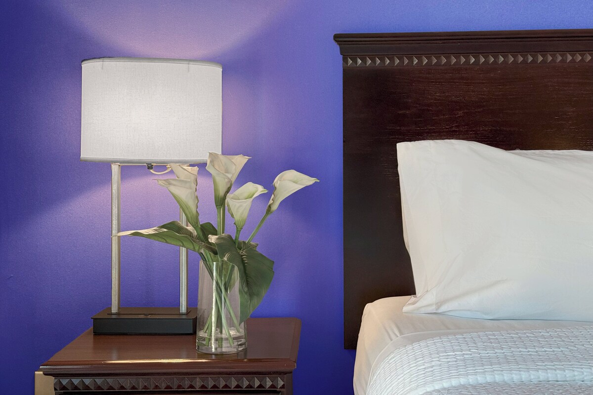 Una cama con cabecera de madera, un almohadón blanco y una mesita de noche con lámpara y un jarrón con flores.