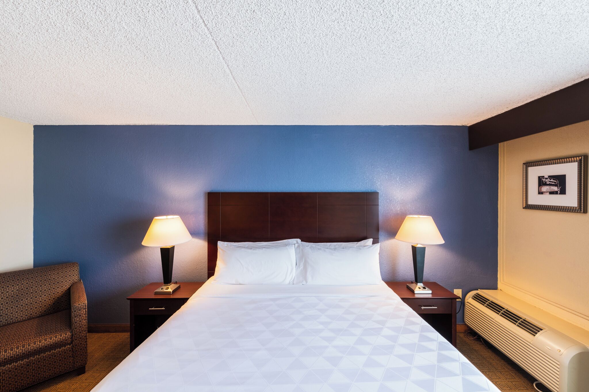 A hotel room with a large bed, two bedside tables with lamps, a chair, and a wall-mounted heater.