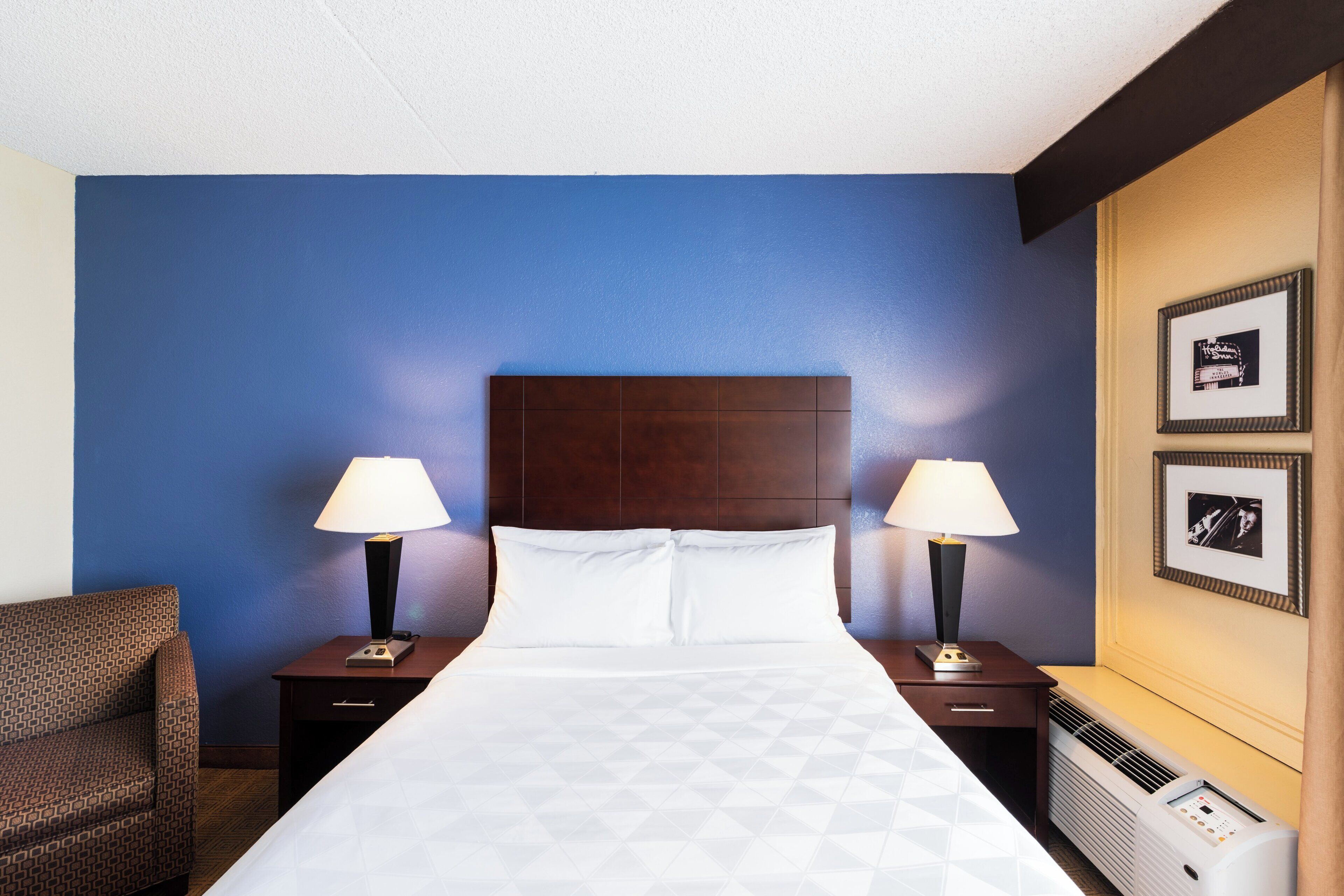 Une chambre d’hôtel avec un grand lit, deux tables de chevet avec des lampes, une chaise et un climatiseur fixé au mur.