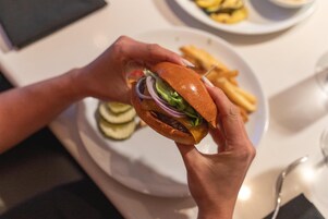 A person holding a burger with visible lettuce, tomato, and onion, and a plate of fries in the background.