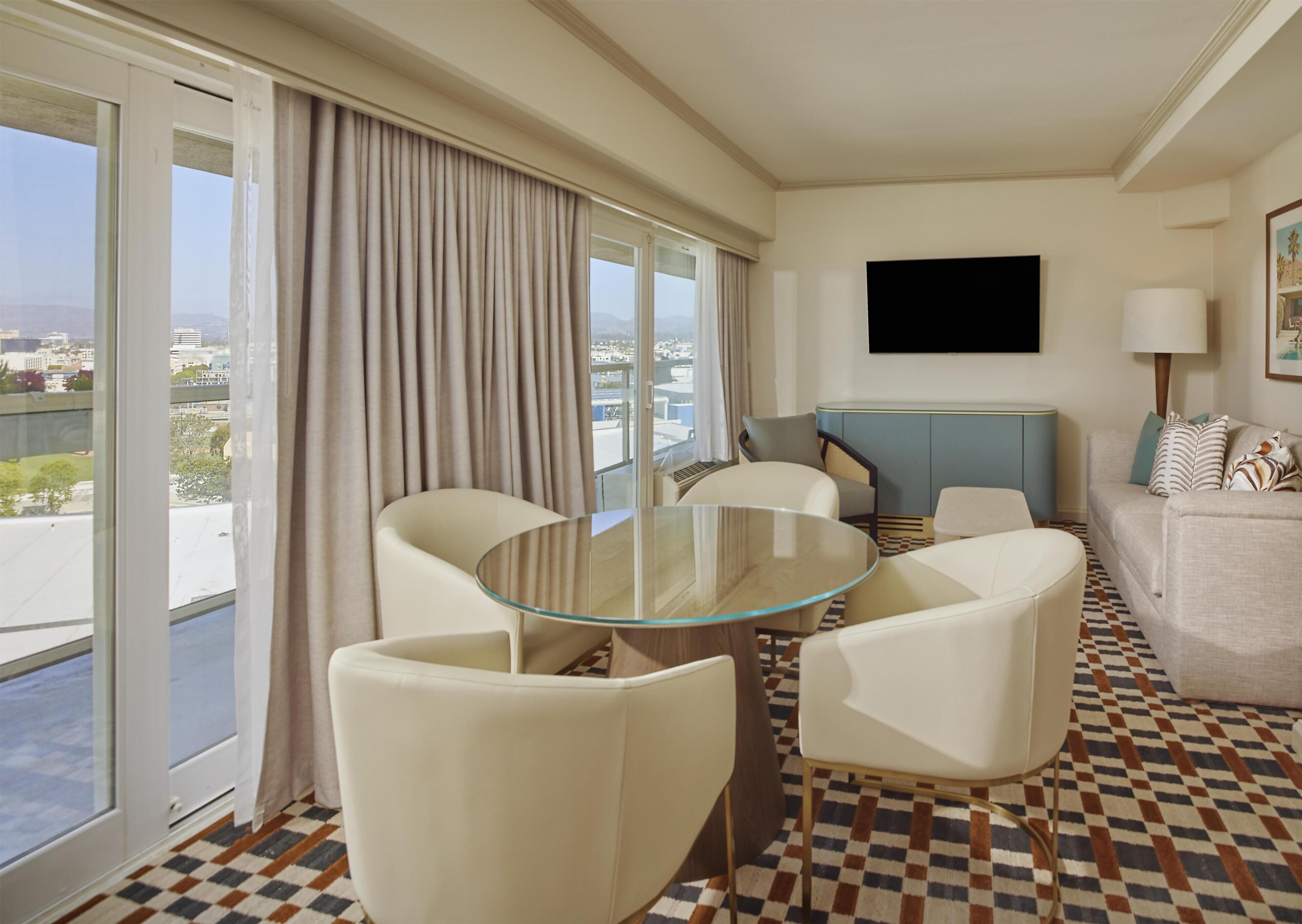 Ein Hotelzimmer mit Glastisch, zwei Stühlen, einer Couch, einem Fernseher und Blick auf die Stadt.
