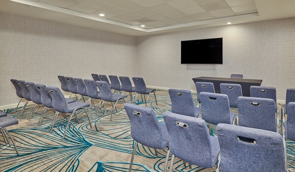 Une salle de conférence avec des rangées de chaises grises, un grand téléviseur à écran plat accroché au mur et une moquette à motifs.