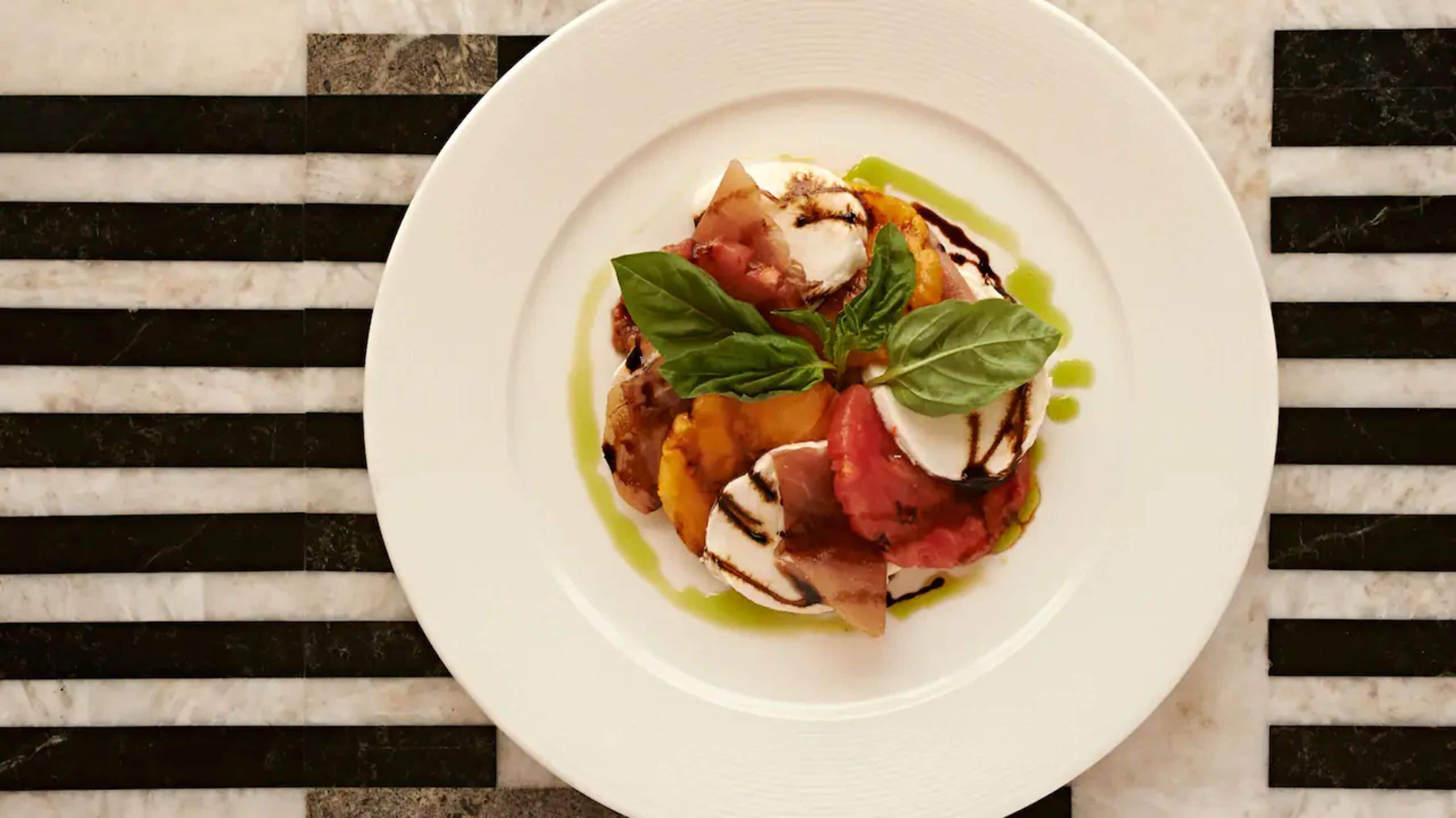 Een wit bord met een portie gegrilde groenten, mozzarella en basilicum op een zwart-wit gestreept oppervlak.