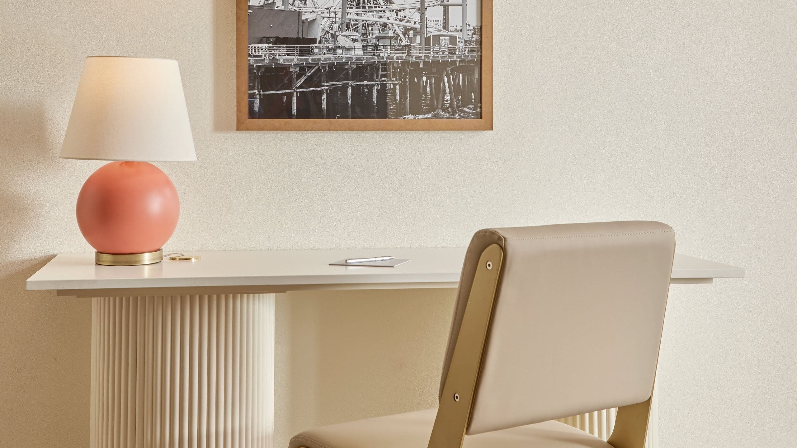 Una habitación con un escritorio blanco, una lámpara rosa, una silla y un cuadro en la pared.