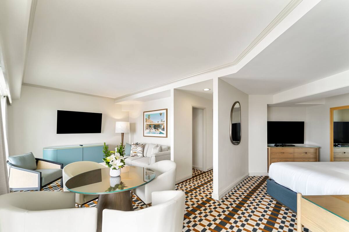 A hotel room with a bed, a seating area with a glass table, a TV, and a painting on the wall.