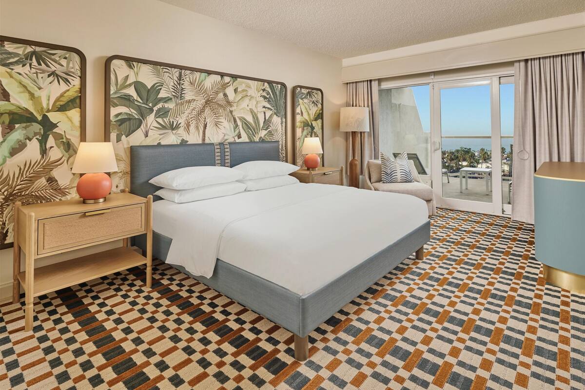 A hotel room with a large bed, two bedside tables with lamps, a view of the outdoors, and a patterned floor.