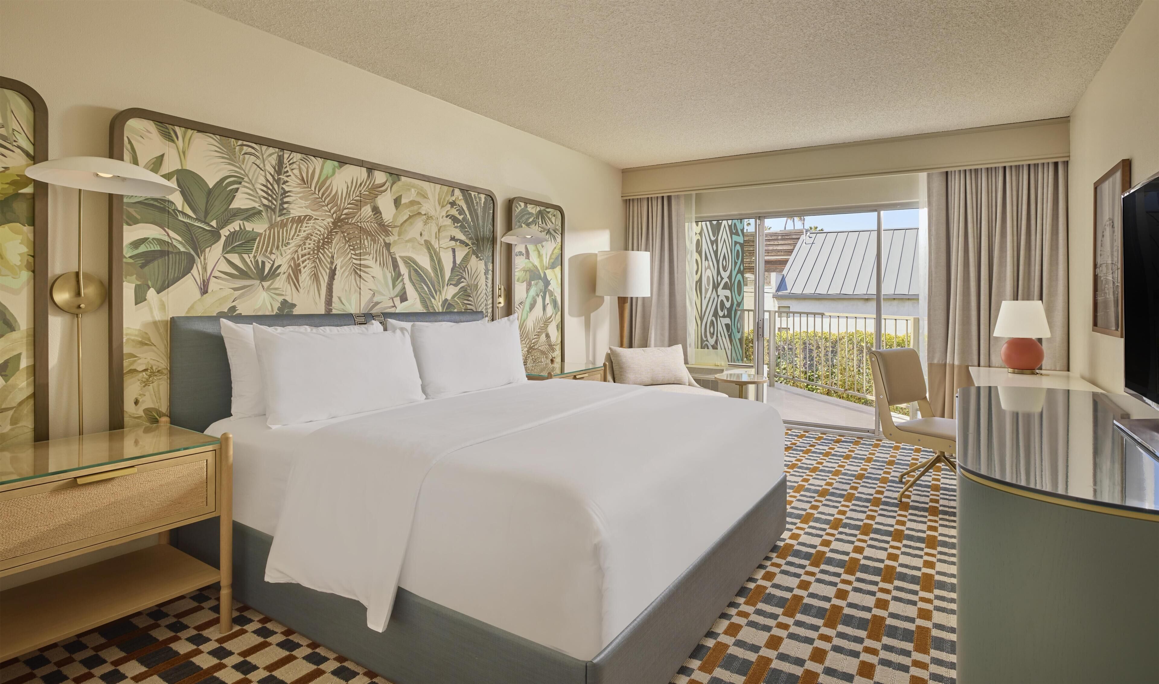 A hotel room with a large bed, a glass side table, a desk with a chair, a TV, and a balcony with a view.