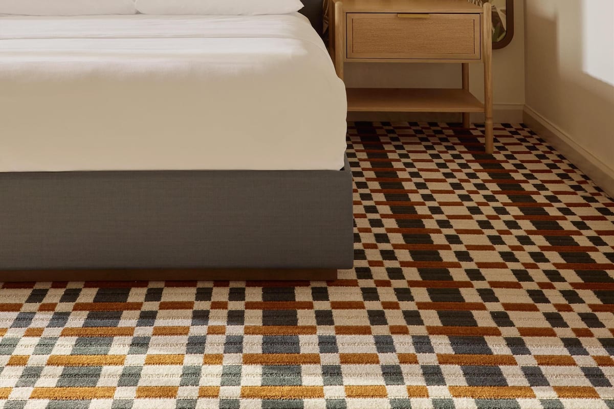 A neatly made bed with a headboard, a bedside table with a lamp, and a patterned floor.