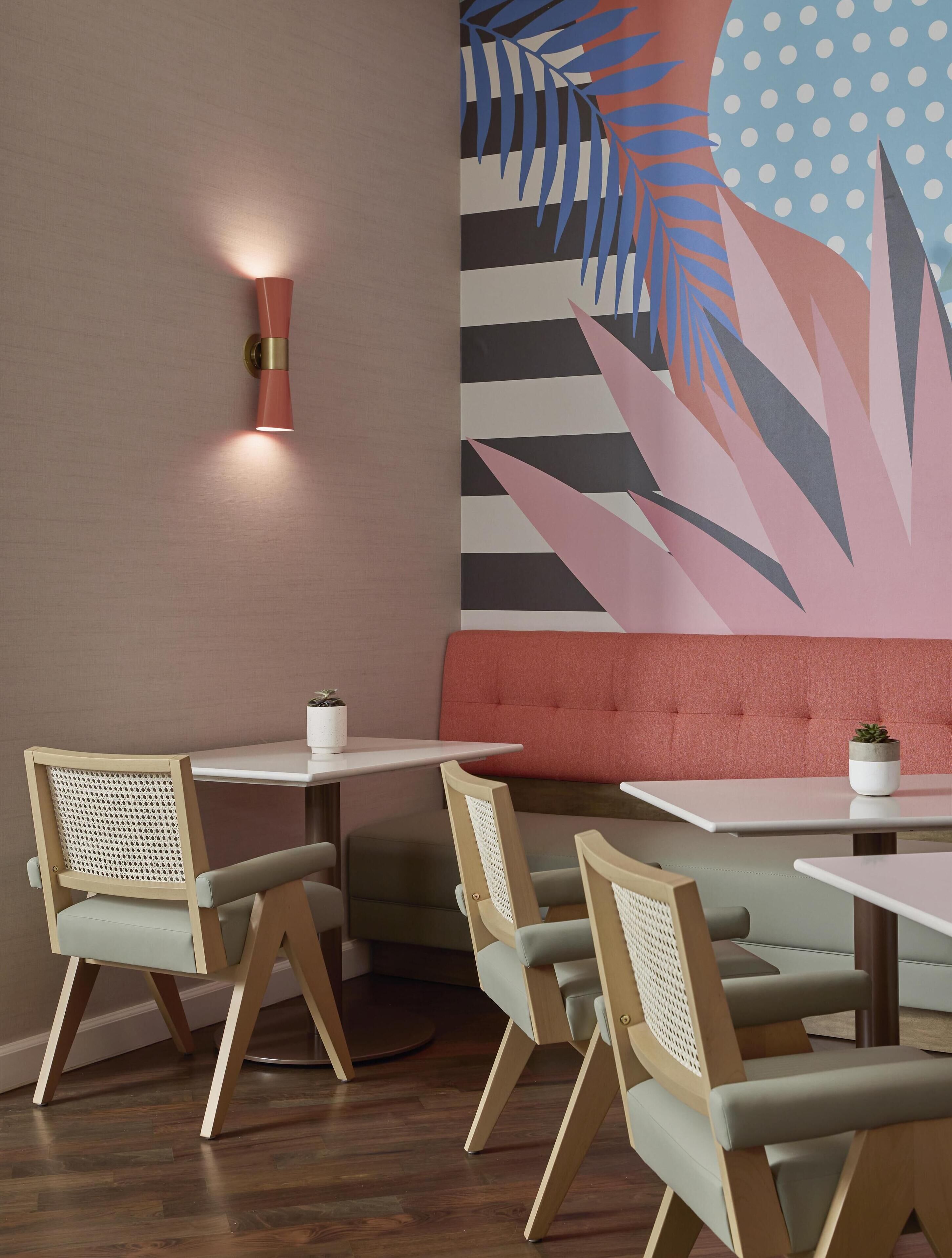 A modern interior with wooden chairs, white tables, a red cushioned bench, and a wall with a tropical leaf pattern.
