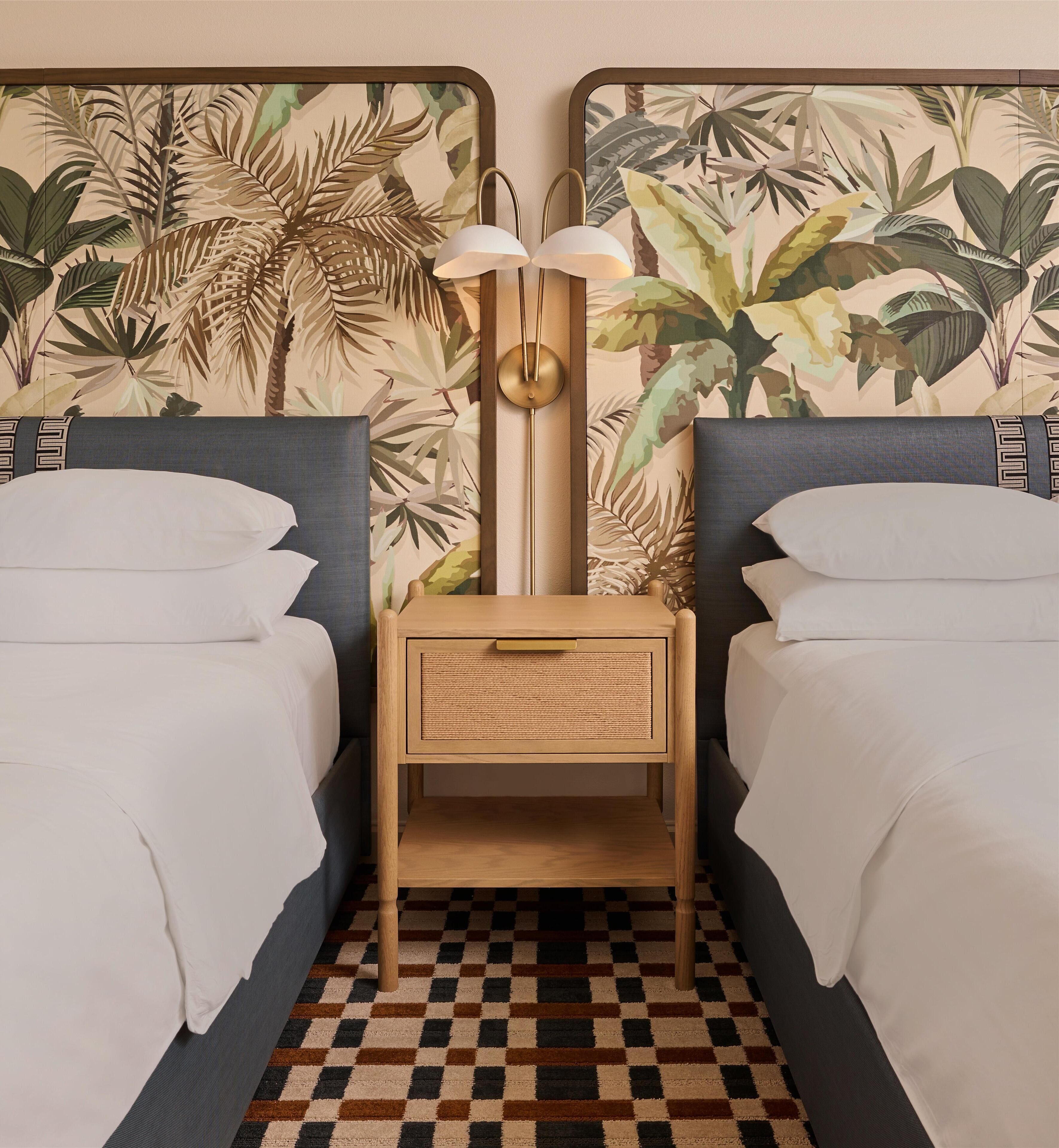 A room with two beds, a nightstand with a lamp, and a wall with tropical leaf wallpaper.