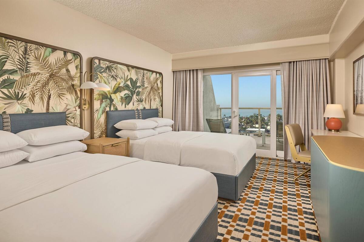 A hotel room with two beds, a desk, a chair, a large window with a view, and a patterned floor.