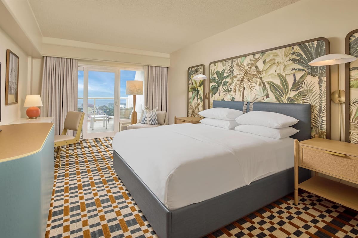 A hotel room with a large bed, a desk, a chair, a balcony with a view, and a tropical wallpaper design.