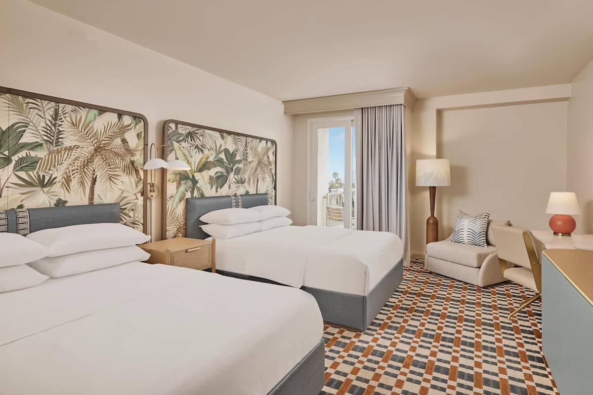 A hotel room with two beds, a desk, a chair, a window with curtains, and a patterned floor.