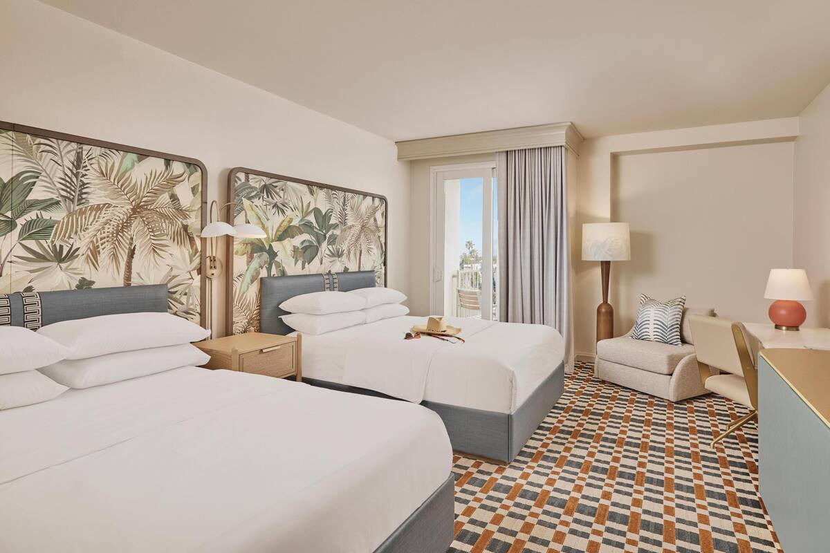 A hotel room with two beds, a desk, a chair, a window with curtains, and a patterned floor.