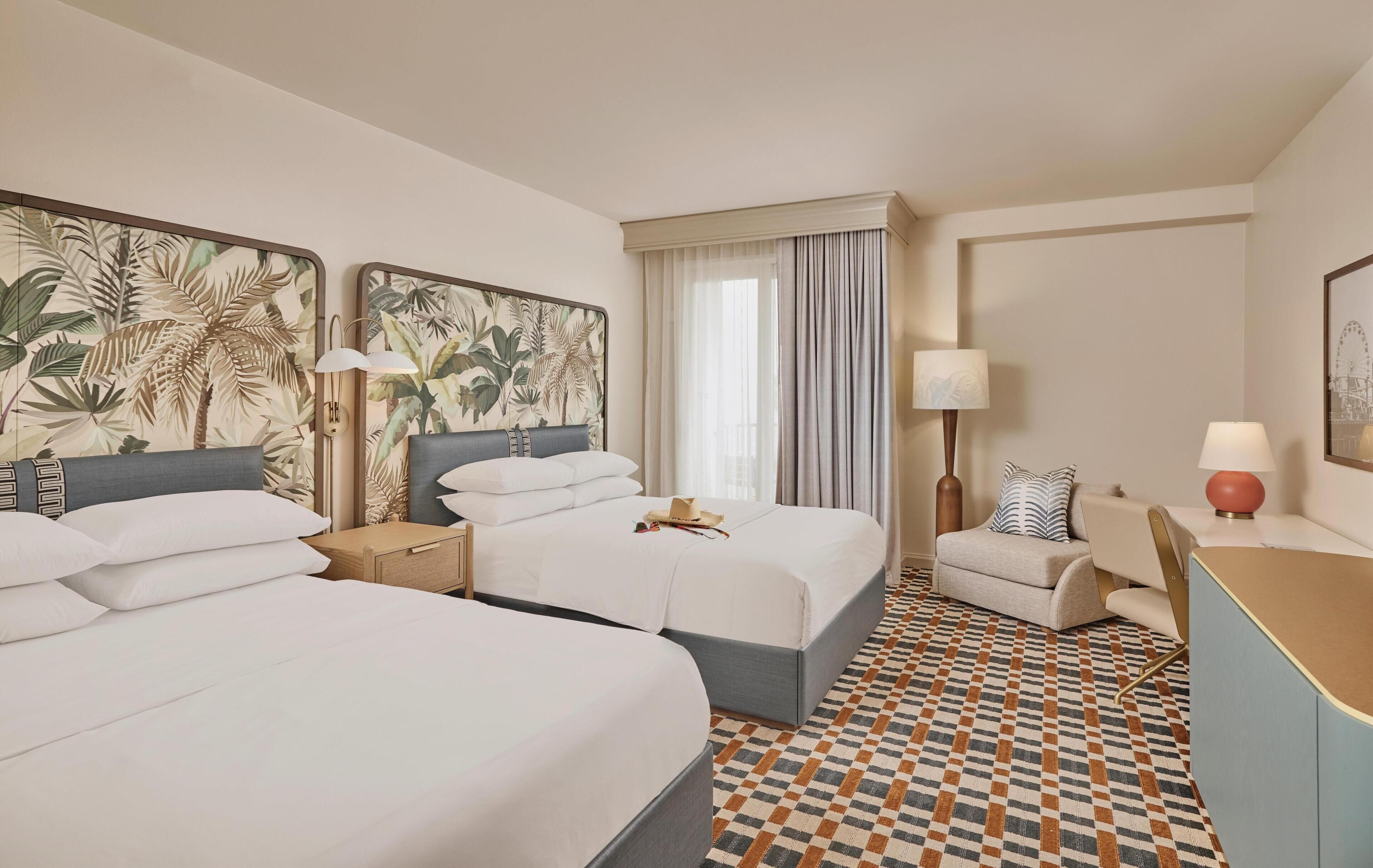A hotel room with two beds, a desk, a chair, a patterned floor, and artwork on the walls.