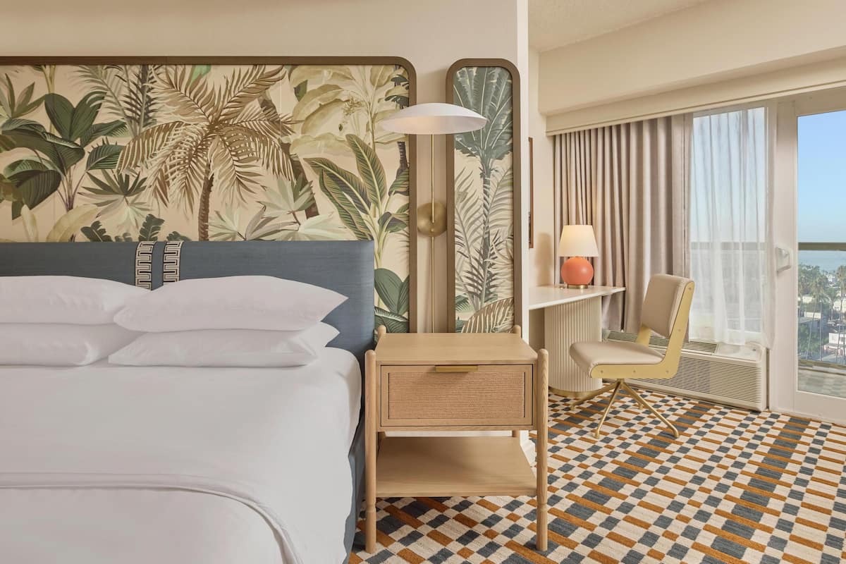 A hotel room with a bed, a nightstand, a chair, a window with a view, and a patterned floor.