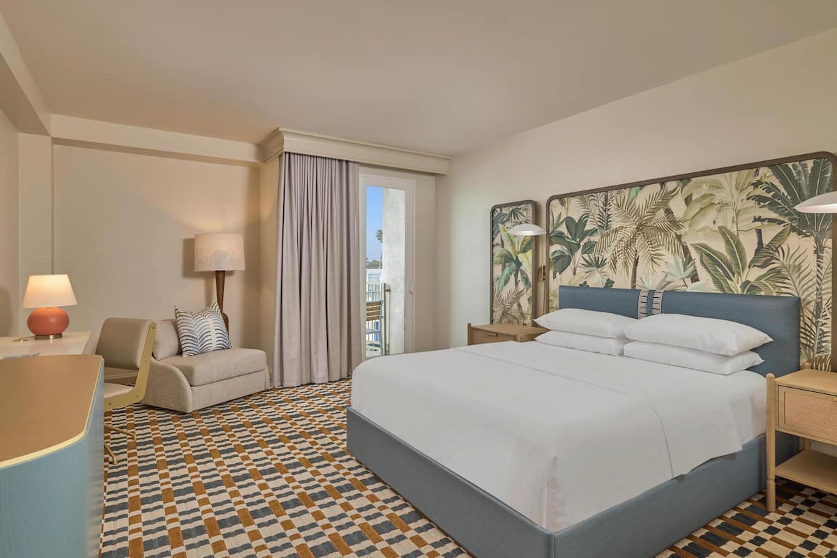 A hotel room with a bed, a desk, a chair, a lamp, and a view of the outside.