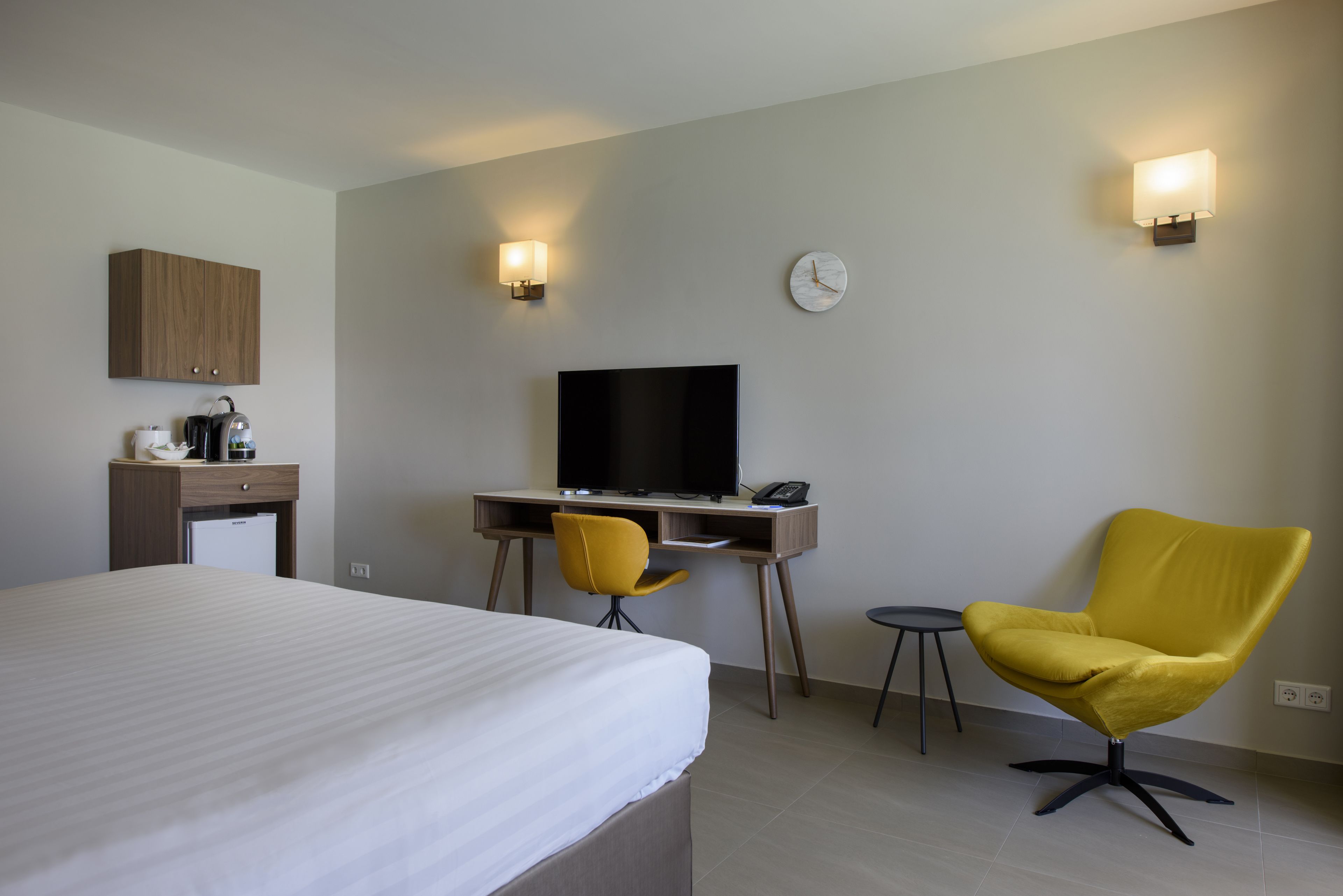 A hotel room with a bed, a desk with a television, a yellow chair, a small table, and a clock on the wall.
