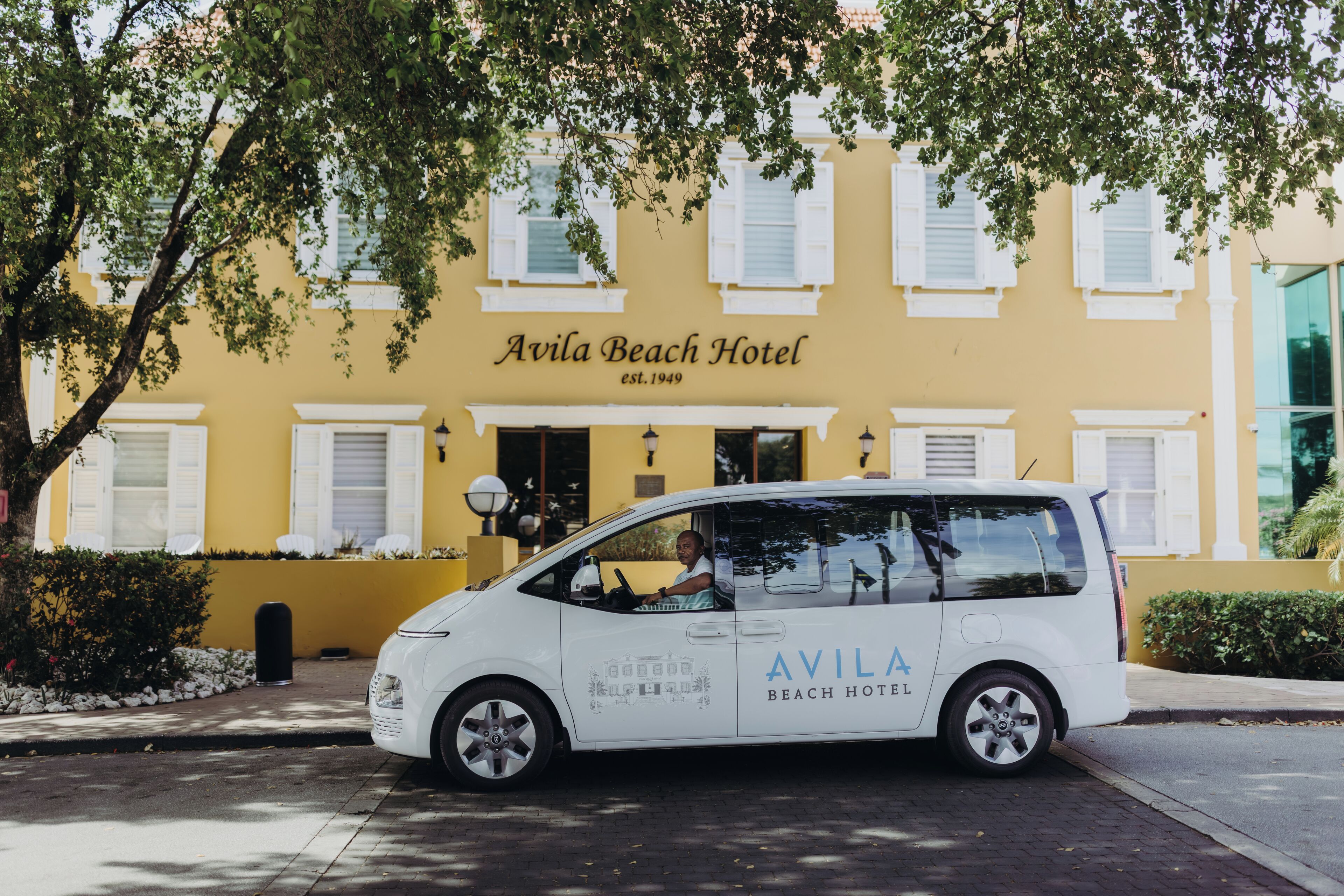 Un coche blanco estacionado frente al Hotel Avila Beach, con fachada amarilla y contraventanas blancas.