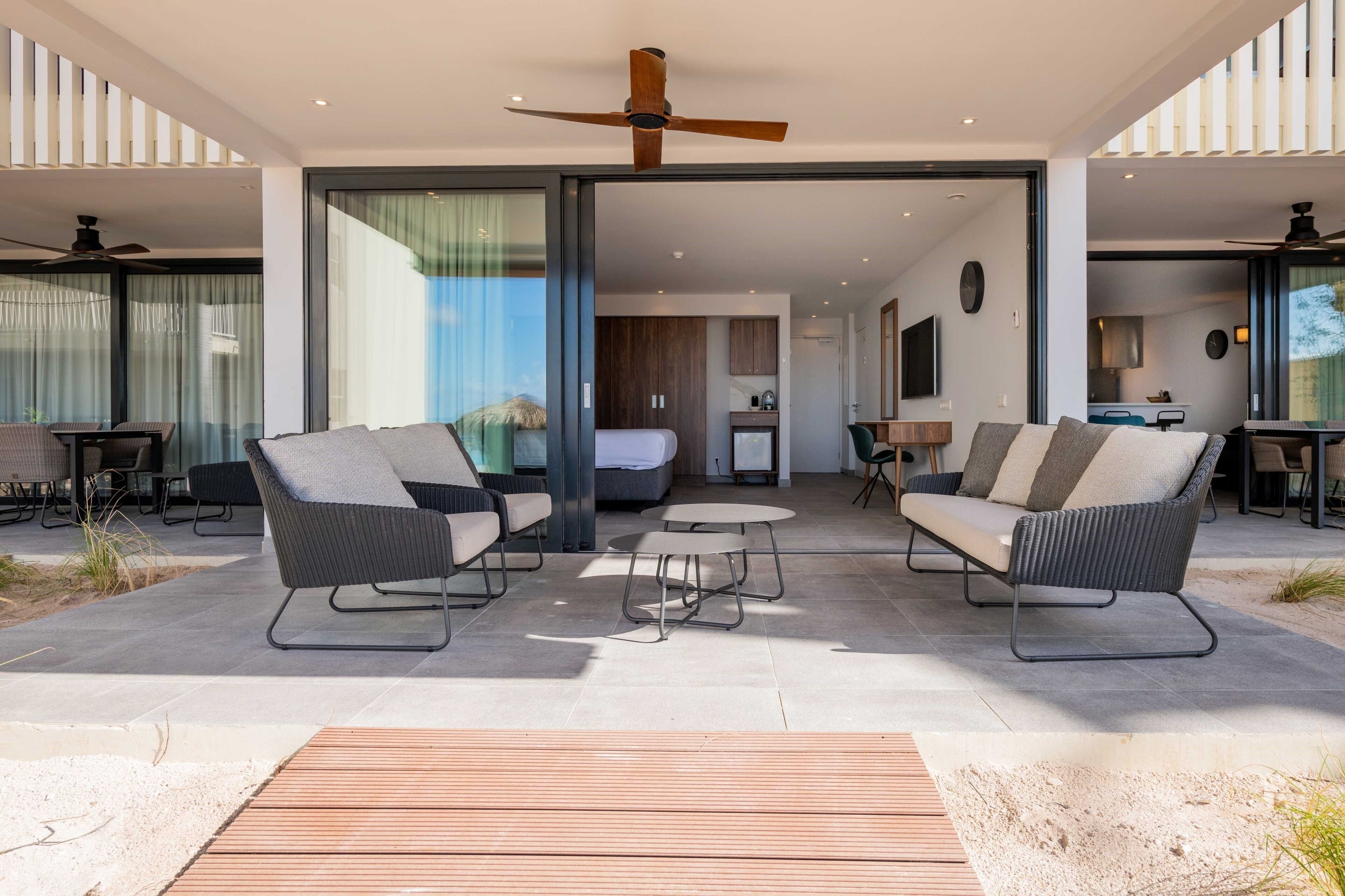 Un espacio moderno al aire libre con dos sofás y una pequeña mesa, conectado a una zona de dormitorio y cocina mediante puertas de cristal.