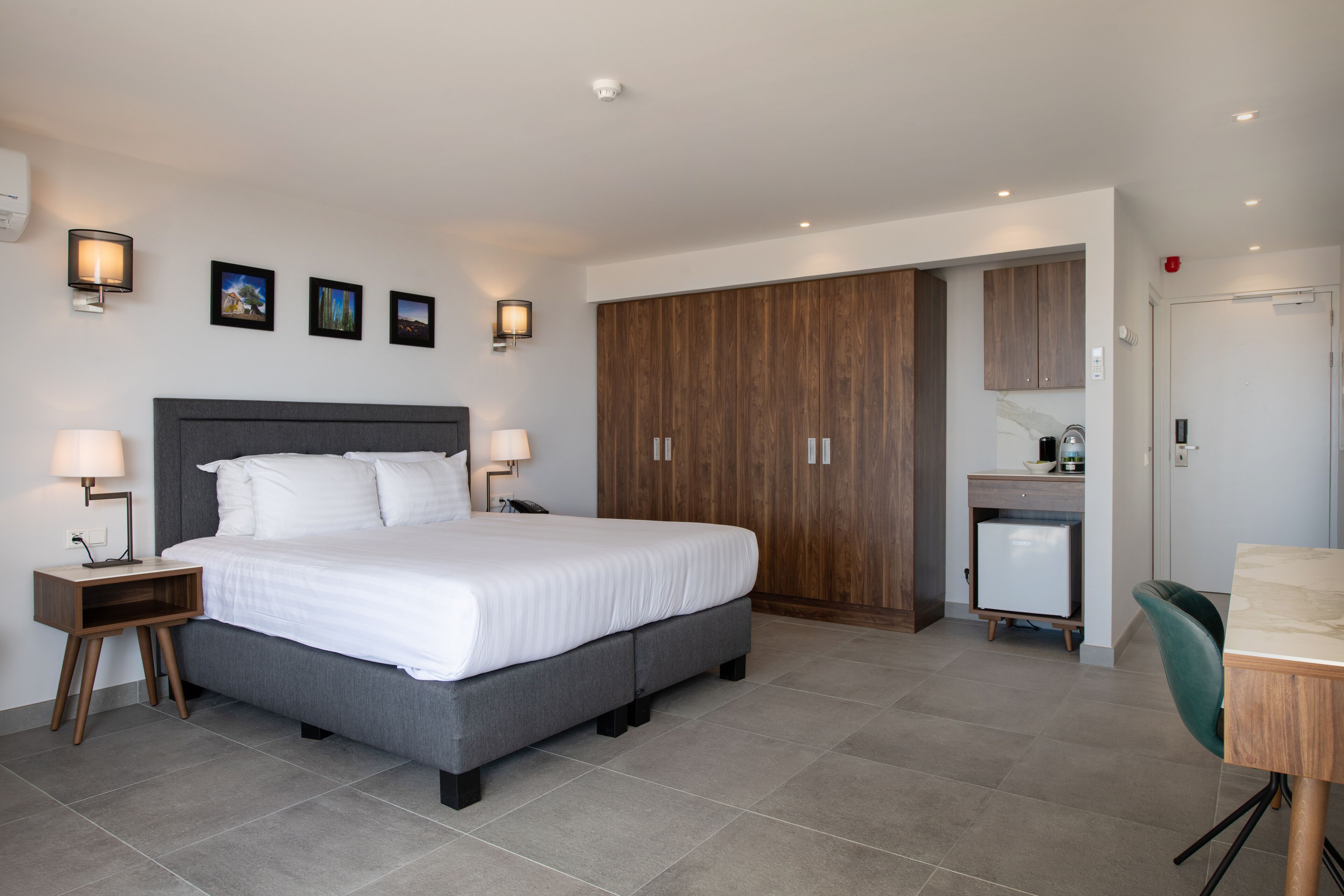 A modern bedroom with a large bed, bedside tables with lamps, a wardrobe, a desk with a chair, and a small kitchenette.