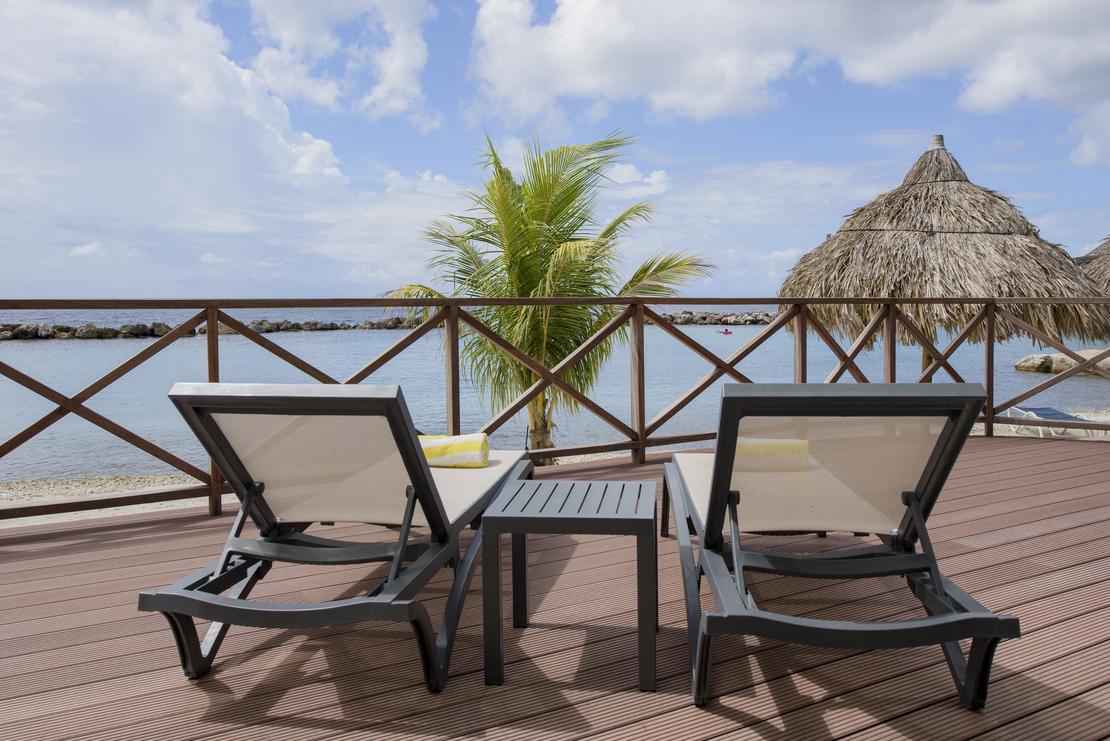 A deck with two lounge chairs and a table, overlooking a body of water with a thatched roof structure.