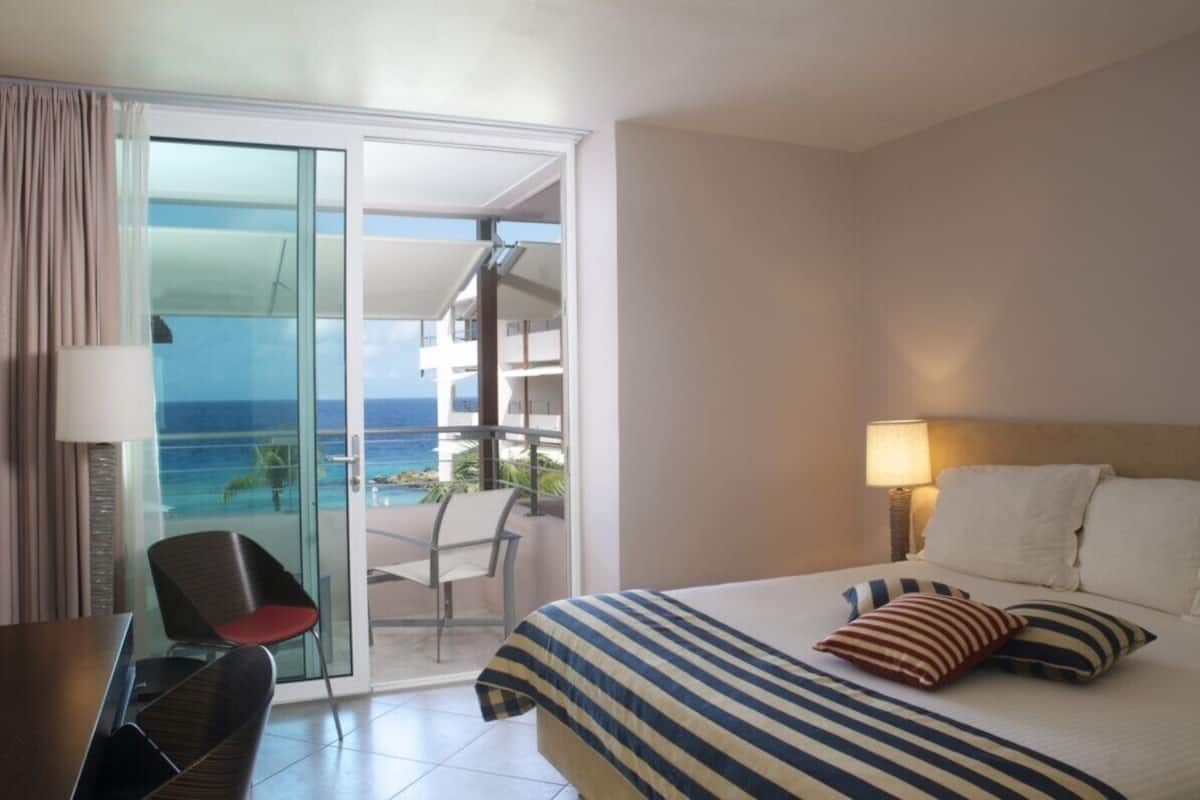 A hotel room with a bed, a chair, a table, and a balcony with a view of the ocean.