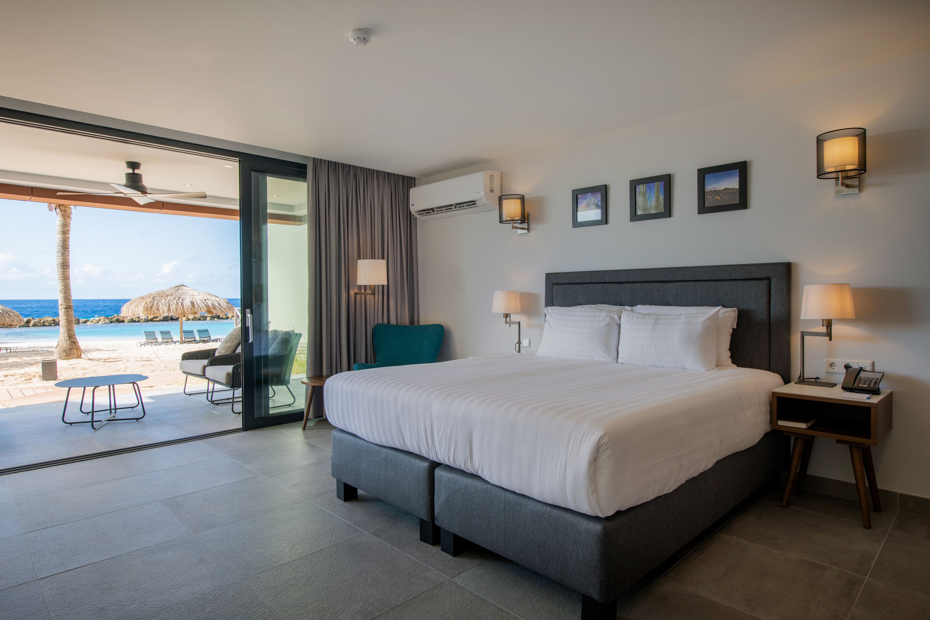 Habitación de hotel con una cama grande, dos mesitas de noche, vista a la playa y un ventilador de techo.