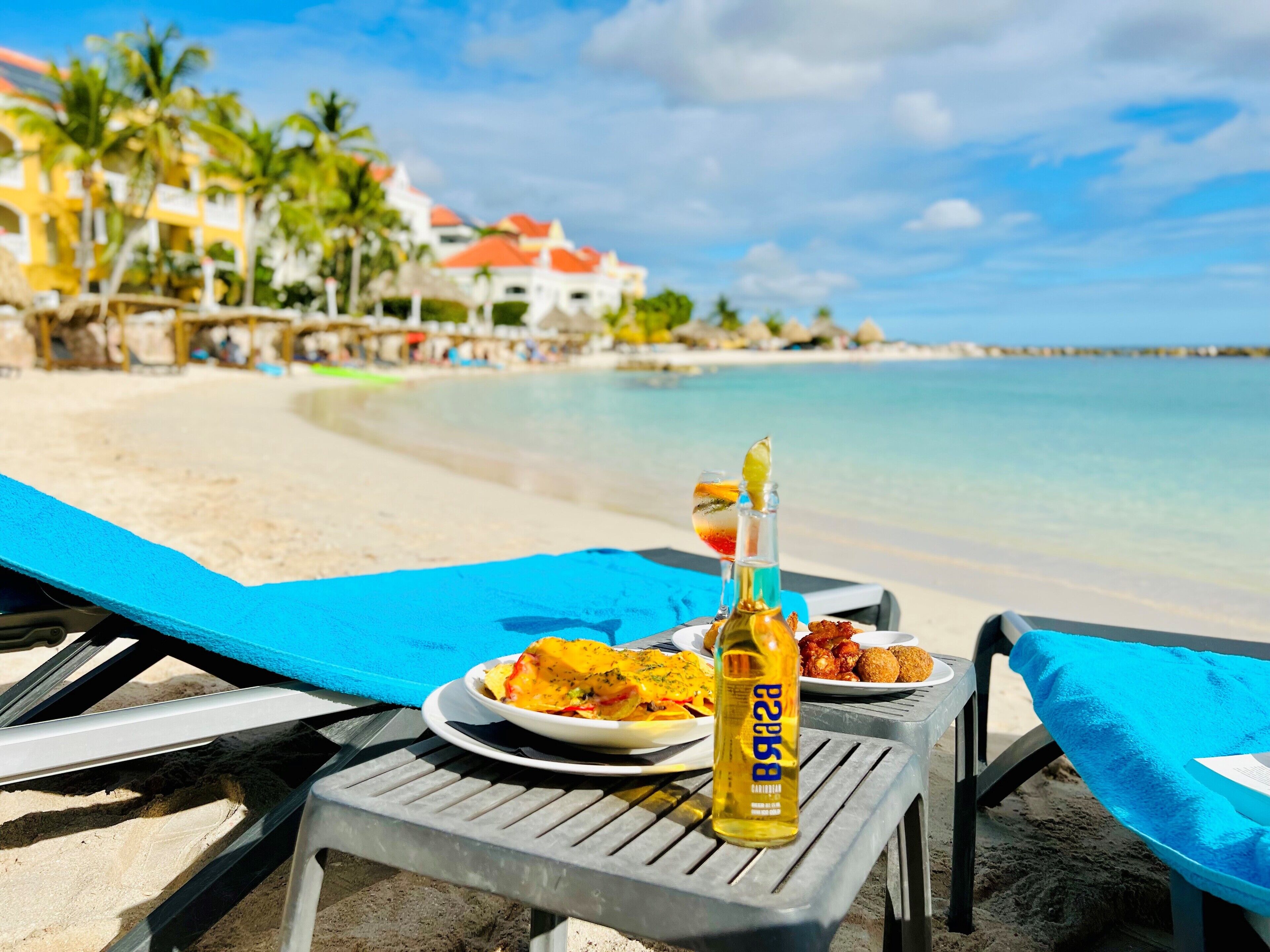 Une table installée sur la plage, avec une bouteille de spiritueux, des assiettes remplies de nourriture et une vue sur l’océan et les bâtiments du complexe hôtelier.