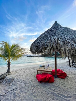 A beach setting with a thatched umbrella, a small palm tree, and a table with red cushions.