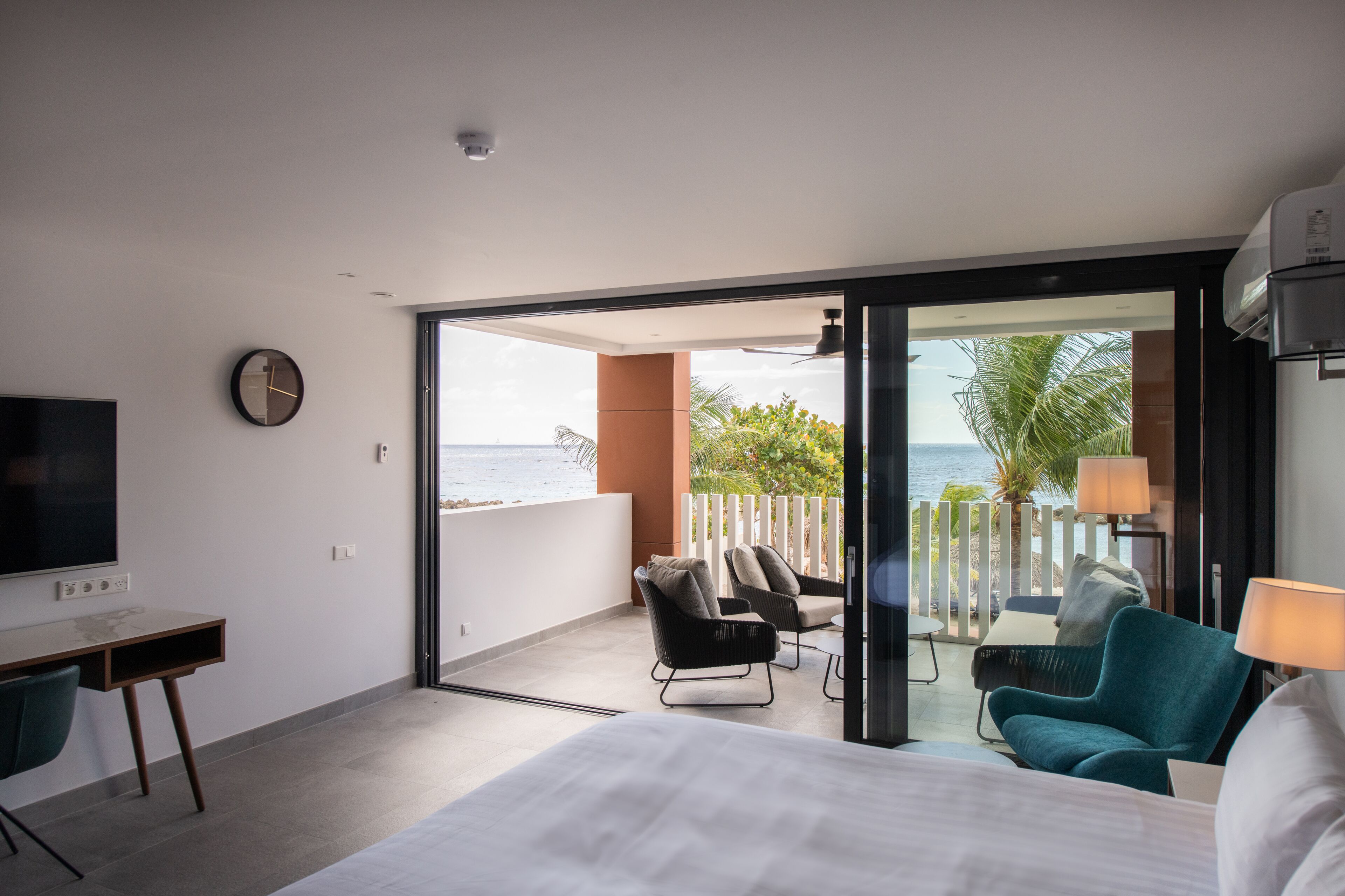 A hotel room with a bed, a desk, a TV, a balcony with chairs, and a view of the ocean.