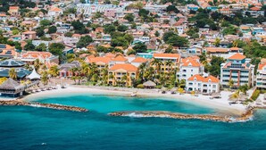 An aerial view of a coastal resort with multiple buildings, a beach, and two clear blue lagoons. - Avila Beach Hotel (Willemstad)