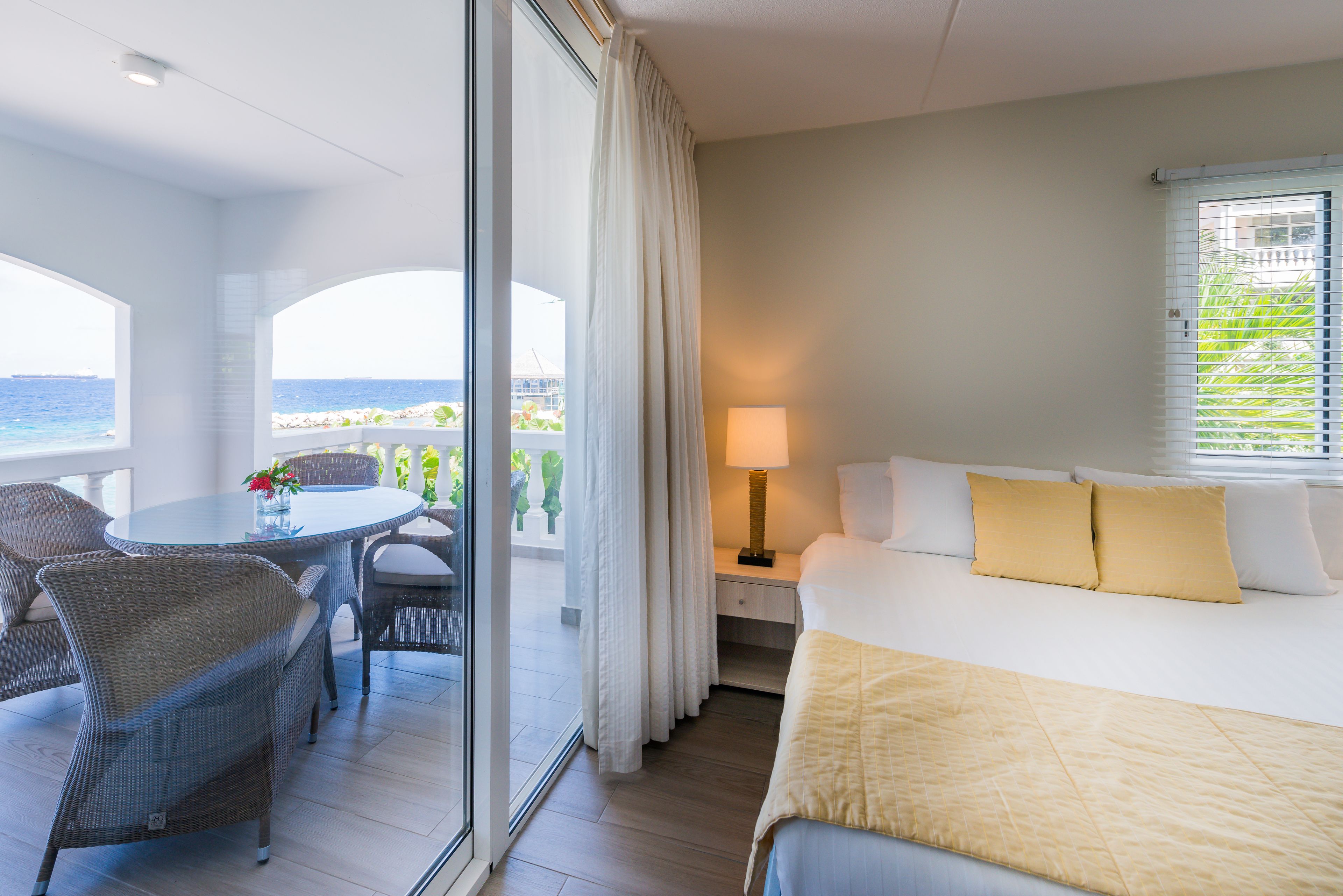 Quarto de hotel com cama, mesa de cabeceira, luminária, mesa com cadeiras e vista para o mar através de grandes janelas.