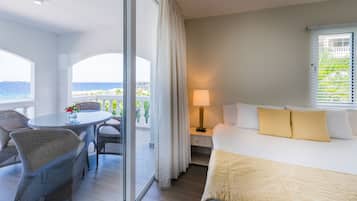A hotel room with a bed, a nightstand, a lamp, a table with chairs, and a view of the sea through large windows.