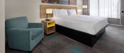 A hotel room with a large bed, a teal armchair, a nightstand with a lamp, and a painting above the bed.
