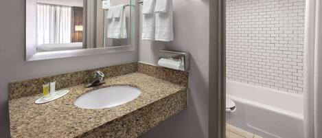 A bathroom with a granite countertop sink, a mirror, a towel rack with two towels, a hair dryer, and a white tiled shower area.