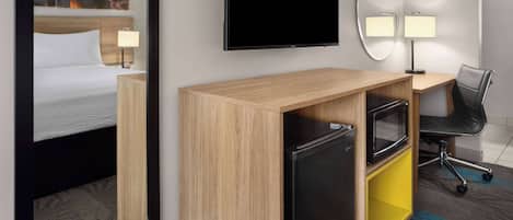 A hotel room with a bed, a desk with a computer, a TV, a mirror, and a chair.