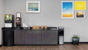 Ein Empfangsbereich mit Kaffeemaschine, Kühlschrank und Mülleimer. An der Wand hängen gerahmte Bilder und es steht ein Blumentopf bereit.