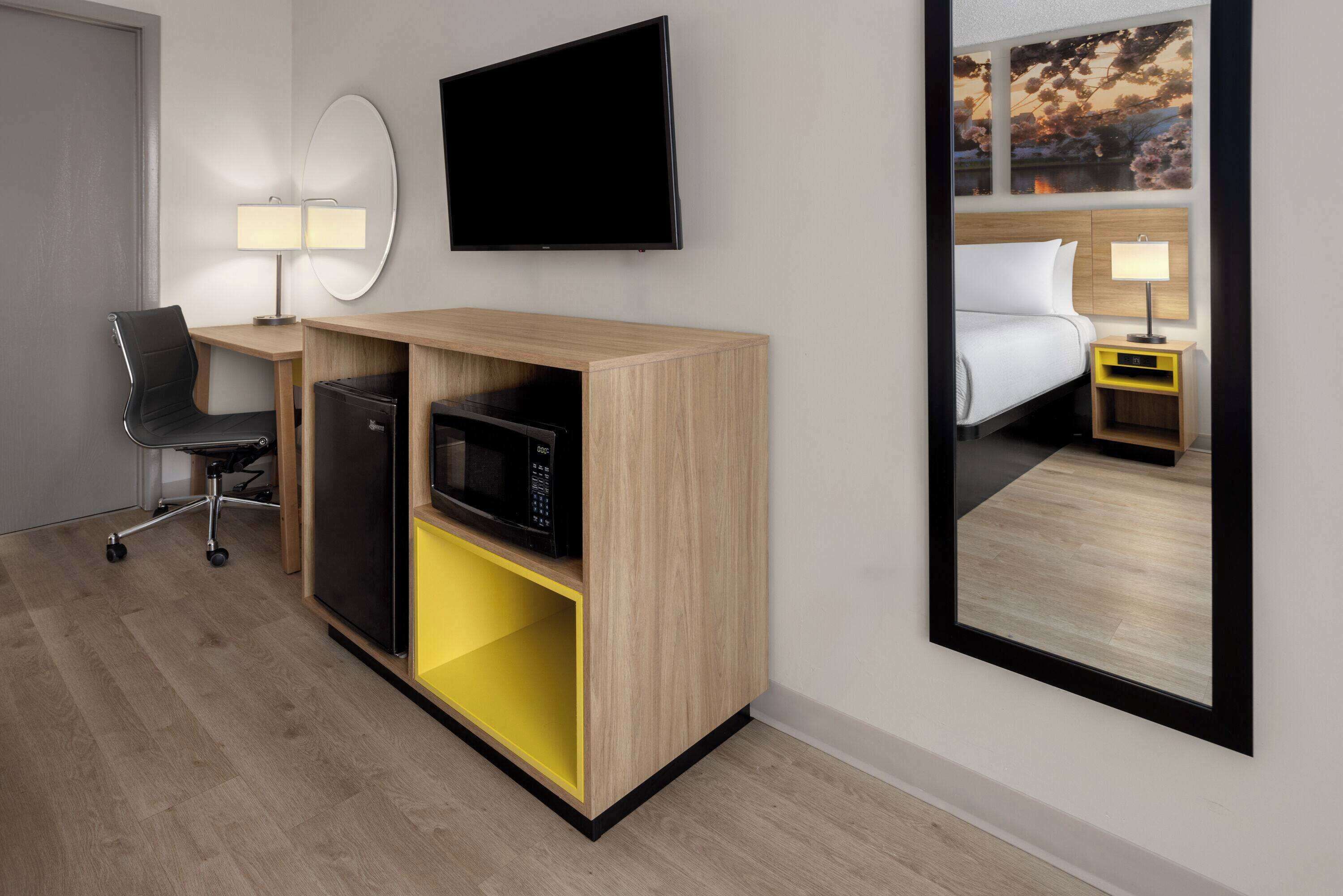 Una habitació d'hotel amb un llit, una escriptori amb una cadira, una televisió, un microones, una taula petita amb una làmpada i un mirall.